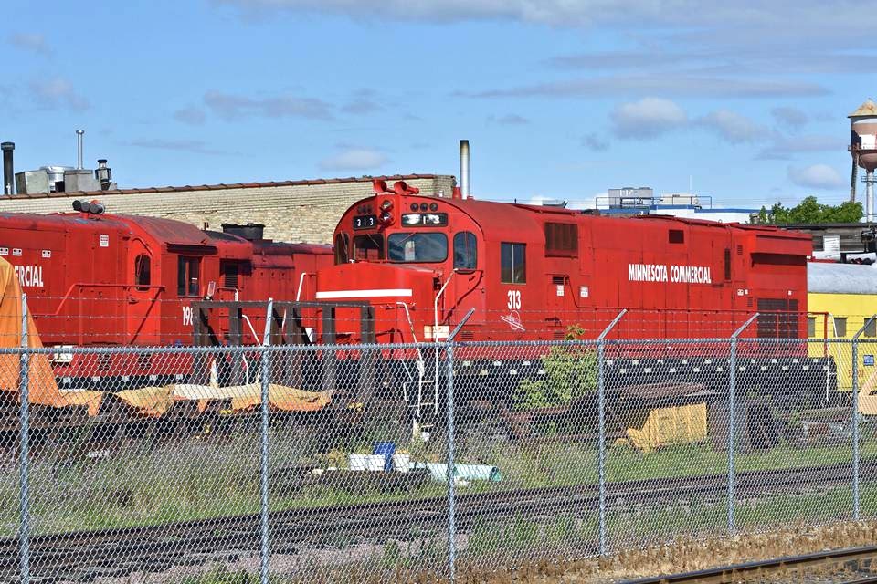 Two locomotives are stopped along the railway tracks surrounded by galvanized chain link fence.