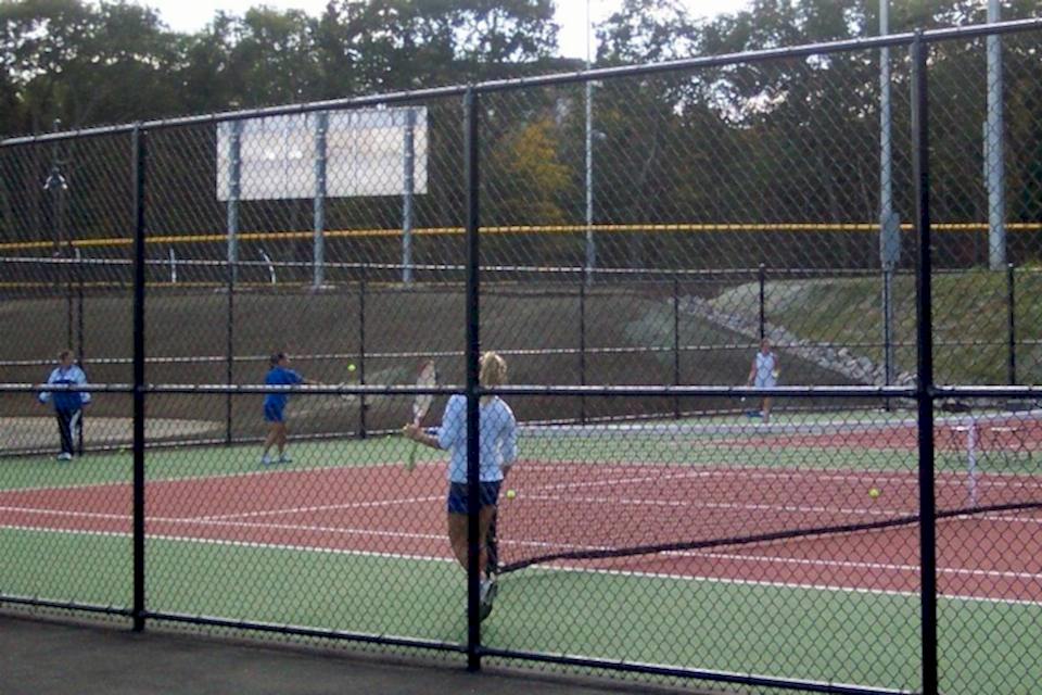 Several players are tennis in the tennis court enclosed by black PVC coated chain link fence.