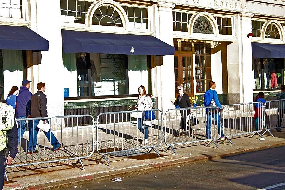 Many people are walking along the commercial street that crowd control barrier is placed.